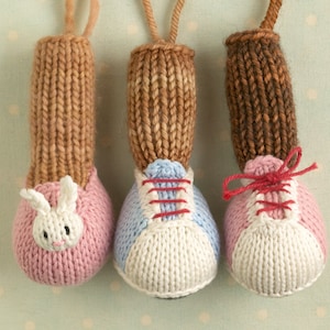 Puede incluir: Tres zapatos de juguete de punto con diferentes diseños. El primer zapato es rosa con una cara de conejo blanco. El segundo zapato es azul y blanco con cordones rojos. El tercer zapato es rosa y blanco con cordones rojos.
