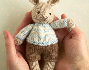 May include: A knitted bunny toy wearing a blue and white striped sweater and brown pants. The bunny is held in two hands.