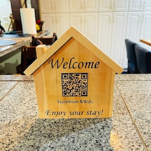 May include: A wooden house-shaped sign with the word "Welcome" and a QR code. The sign also says "Guidebook & Wi-Fi" and "Enjoy your stay!"