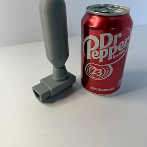 May include: A gray, 3D-printed object with a cylindrical handle and a rectangular base, next to a red Dr. Pepper can. The can is 12 fl oz (355 ml).