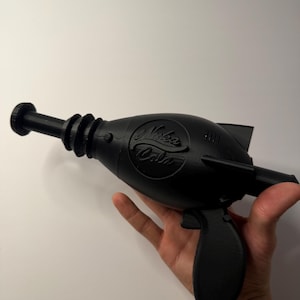 May include: Black toy ray gun with the "Nuka Cola" logo. The gun has a long barrel, a trigger, and a handle. The ray gun is held in a hand against a white background.