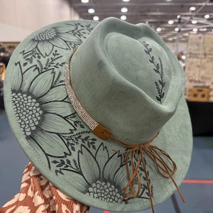 Puede incluir: Sombrero fedora verde salvia con diseño de girasoles y hojas negras. El sombrero presenta una banda de pedrería, una correa de cuero marrón y un lazo marrón. Un accesorio elegante.