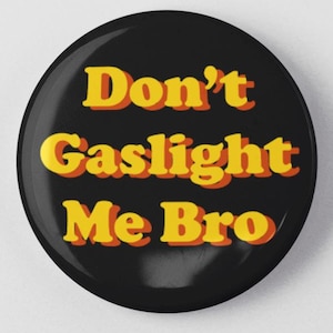 May include: A round, black button with the text "Don't Gaslight Me Bro" in yellow and orange lettering. The button is centered on a white background. The text has a retro, bold font.