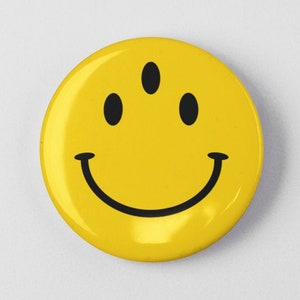 May include: A yellow smiley face button with three black eyes and a black smile.