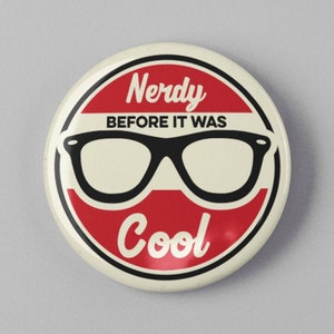 May include: A round button with a red and white design. The button features the text "Nerdy BEFORE IT WAS Cool" with a pair of black glasses in the center. The button is on a gray background.
