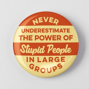 May include: A round button with a cream and orange color scheme. The button features the text: "NEVER UNDERESTIMATE THE POWER OF STUPID PEOPLE IN LARGE GROUPS" in a bold, vintage font.