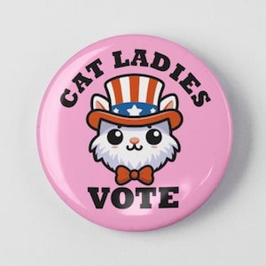 May include: A pink button with a white cat wearing a red, white, and blue Uncle Sam hat. The text "Cat Ladies Vote" is written around the cat.