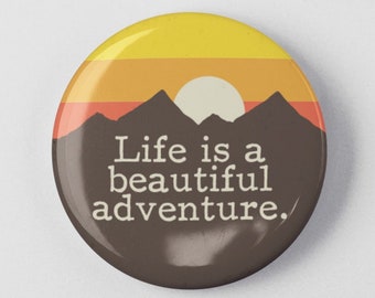 Pode incluir: Um botão marrom com um gráfico de pôr do sol e o texto "Life is a beautiful adventure."