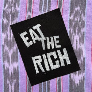 May include: A rectangular black fabric patch featuring the white embroidered text EAT THE RICH in a bold, sans-serif font. The patch has a visible woven texture and clean, straight edges, presenting a high-contrast design with clear, legible lettering.
