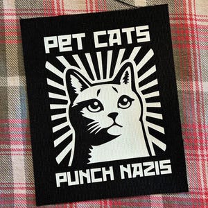 May include: Black patch with white text and a cat illustration. The text reads "PET CATS" above the cat's head and "PUNCH NAZIS" below. The cat is in the center, surrounded by radiating lines.