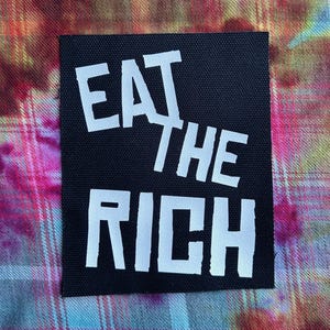May include: Black rectangular patch with the white text "EAT THE RICH". The patch is on a colorful, patterned fabric background. The text is in a bold, sans-serif font.
