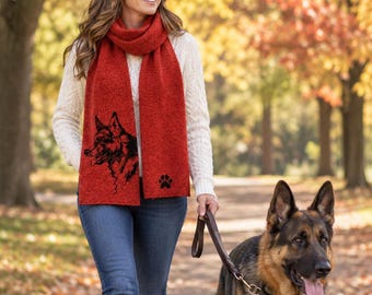 May include: A vibrant red knit scarf featuring a black graphic illustration of a German Shepherd dog profile and a small black paw print icon. The scarf has a textured, woven appearance and is worn draped around the neck over a white sweater.