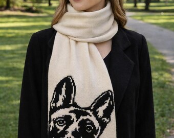 May include: Cream-colored scarf featuring a black and white dog face design. The scarf is worn over a black coat. The dog's face is prominently displayed on the scarf, adding a unique touch to the accessory. The scarf is made of a soft material.