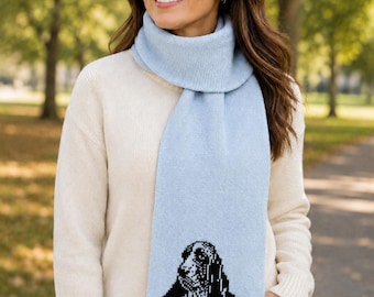 May include: A light blue knit scarf featuring a black and white graphic illustration of a Basset Hound dog. The scarf is made of a soft, textured material and is worn draped around the neck, showcasing the detailed canine portrait at the bottom end.