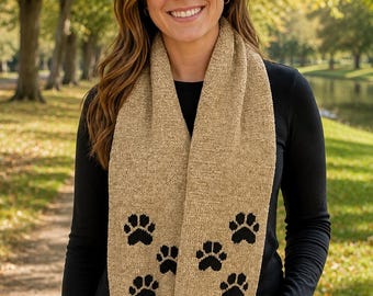 May include: A tan knit scarf featuring a repeating pattern of black paw prints. The scarf has a textured fabric construction and is finished with two black pom-poms at the bottom ends, providing a decorative accent to the long, rectangular accessory.
