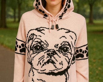 May include: A light pink hooded sweater featuring a large black pug face design on the front. The hood has a black and white spotted pattern. Black paw prints are on the sleeves. The sweater is made of a soft knit material.