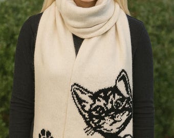 May include: Cream-colored knitted scarf featuring a black cat design and paw print. The scarf is wrapped around the neck and drapes down the front. The cat design is on the lower right side of the scarf.