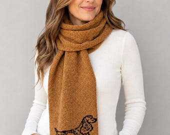 May include: A long, textured, caramel-brown knit scarf featuring a black embroidered outline of a spaniel dog at the bottom end. The scarf is wrapped around the neck, with the ends hanging down to display the detailed canine silhouette design.