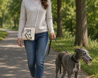 May include: A cream-colored crossbody bag featuring a black line-art illustration of a greyhound dog head. The bag has a thin, matching cream-colored shoulder strap and a rectangular shape, worn across the torso with a simple, clean design.