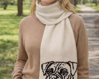 May include: Cream-colored scarf with a black Boxer dog face design. The scarf is draped over a tan sweater. The woman is wearing blue jeans and is standing outside on a path.
