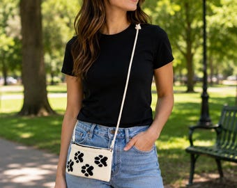 May include: A small, rectangular, off-white crossbody bag featuring a repeating black paw print pattern. The bag has a black zipper closure along the top edge and is attached to a thin, white rope-style shoulder strap with a knotted detail.