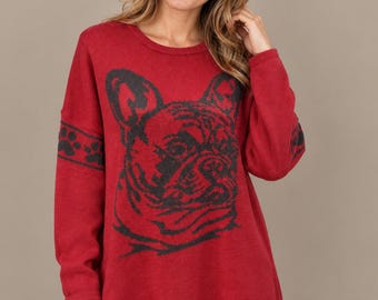 May include: A long-sleeved red knit sweater featuring a large black graphic of a French Bulldog face on the front and a black paw print pattern on the sleeves. The sweater has a round neckline, a relaxed fit, and small side slits at the hem.