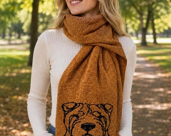May include: A mustard yellow knit scarf featuring a black line art illustration of a Shar-Pei dog face. The scarf has a textured, ribbed appearance and is draped around the neck, showcasing the detailed canine portrait on the lower section.