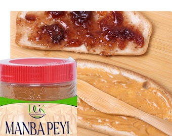 May include: A jar of peanut butter labelled "MANBA PEYI HOT" with a red lid. The jar is on a wooden surface with two slices of bread. One slice of bread is topped with red jelly and the other slice is topped with peanut butter.