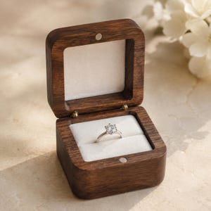 May include: A square dark brown wooden ring box with a hinged lid and a magnetic closure. The interior is lined with soft white fabric, featuring a central slot holding a silver-toned ring with a single round clear gemstone set in a six-prong setting.