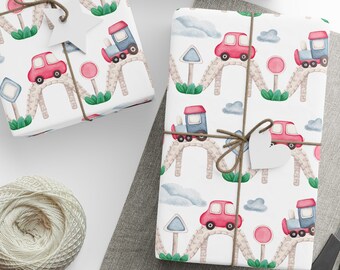 May include: White wrapping paper with a repeating pattern of red cars and blue trucks driving over a bridge. The paper is tied with twine and a white heart-shaped tag.