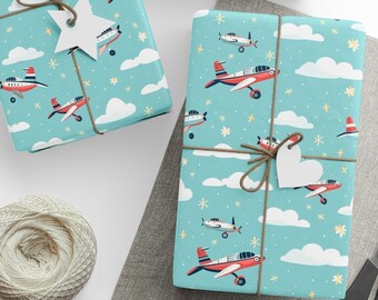 May include: Three blue gift boxes with a white and red airplane pattern. The boxes are wrapped with twine and have white paper tags attached. The boxes are on a gray surface with white paper circles and a pair of scissors.
