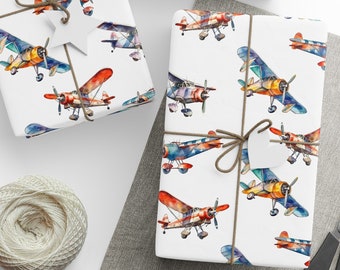 May include: White gift wrap with a repeating pattern of colorful watercolor airplanes. The paper is wrapped around a box and tied with twine. There are also some round white paper circles on a gray surface.