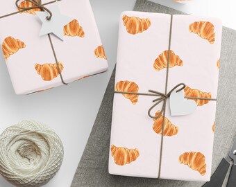 May include: Three gift boxes wrapped in white paper with a brown string and a brown bow. The paper has a pattern of orange croissants. The boxes are on a white surface with a gray fabric surface in the background.