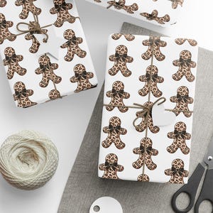 May include: Several wrapped gifts with white paper featuring a repeating pattern of gingerbread men with leopard print bodies and black bows. The gifts are tied with twine and have blank tags. A ball of twine, scissors, and blank tags are also present.