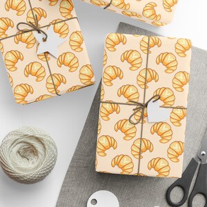 May include: Gift-wrapped boxes with a repeating pattern of cartoon croissants on a beige background. The gifts are tied with twine and have blank white tags. A ball of twine, scissors, and additional tags are also visible.