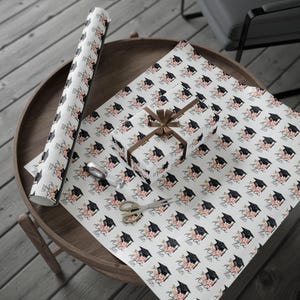 May include: White gift wrap with a repeating pattern of black graduation caps and pink flowers with the text "Class of" printed on each cap.