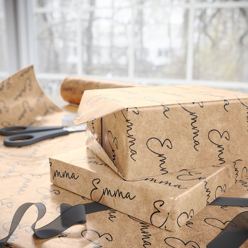 May include: Two stacked gift boxes wrapped in brown paper with the name "Emma" printed in a repeating pattern. A partially unwrapped box reveals the contents. Black scissors and a roll of paper are visible.
