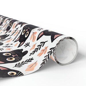 K&ouml;nnte beinhalten: Eine Rolle Geschenkpapier mit einem sich wiederholenden Muster aus schwarzen Katzenk&ouml;pfen, pfirsichfarbenen Bl&auml;ttern und schwarzen Zweigen auf wei&szlig;em Hintergrund. Die Katzen haben gelbe Augen und rosa Innenohren. Das Papier ist aufgerollt und zeigt das Muster auf der Au&szlig;enseite.