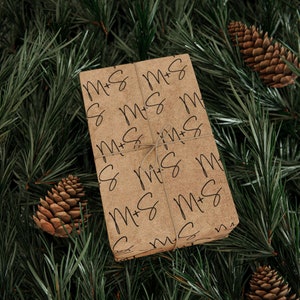 May include: A wrapped gift with a brown paper cover featuring the handwritten letters "M + S" repeated across the surface. The gift is tied with twine and rests on a bed of green pine needles, with several pine cones scattered around.