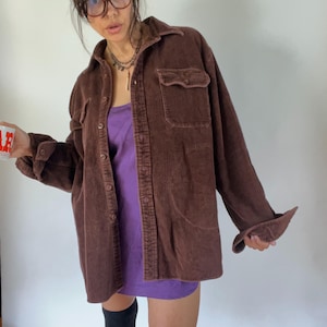 May include: A brown corduroy shirt with two chest pockets and button closures. The shirt is worn over a purple dress. The sleeves are cuffed at the wrists. The shirt has a collared neckline.