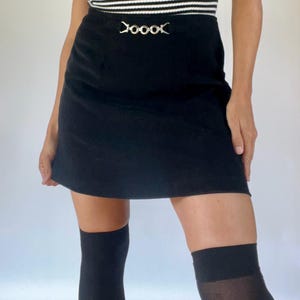 May include: A black mini skirt with a decorative silver-tone buckle. The skirt is made of a smooth fabric and has a classic A-line shape. The model is wearing black knee-high socks and a striped top.