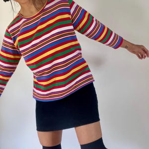 May include: A colorful striped long-sleeve shirt with a round neckline. The shirt features horizontal stripes in red, yellow, green, blue, and white. The shirt is paired with a black mini skirt.