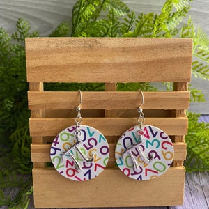May include: A pair of silver earrings with a white background featuring a colorful pattern of numbers. The earrings are shaped like a circle with a silver pi symbol hanging from the bottom.