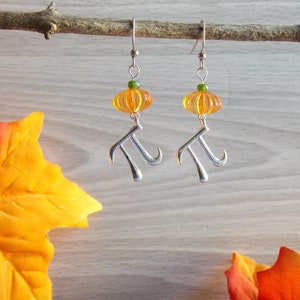 May include: A pair of silver earrings with a pumpkin-shaped bead and a pi symbol charm. The beads are orange and the charms are silver.