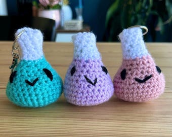 May include: Three crochet keychains shaped like laboratory flasks, each featuring a white top section and a rounded base in teal, light purple, or light pink. Each flask has two black embroidered eyes and a small black embroidered mouth.
