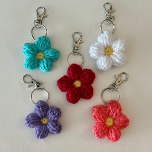 Puede incluir: Cinco llaveros de flores de ganchillo en diferentes colores: turquesa, blanco, rojo, morado y coral. Cada flor tiene un centro amarillo y está unida a un llavero plateado.