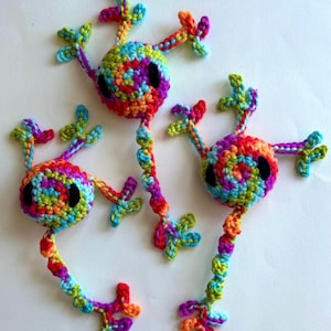 Può includere: Tre creature fatte a mano all'uncinetto in vivaci colori arcobaleno. Ognuna ha una testa rotonda con occhi neri, un corpo lungo e strutturato e molteplici appendici multicolori. Le creature sono esposte su uno sfondo bianco.