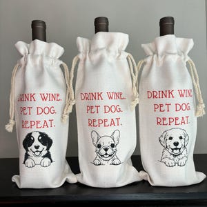 May include: Three white canvas wine bags with red text and black dog illustrations. The text on the bags reads "Drink Wine. Pet Dog. Repeat." Each bag features a different dog breed: a Bernese Mountain Dog, a French Bulldog, and a Golden Retriever.