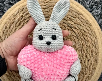 May include: A gray crocheted bunny holding a pink heart-shaped plush toy.