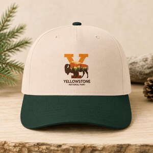May include: A cream-colored baseball cap with a dark green brim. The front features a graphic of a large orange letter Y containing a silhouette of a bison and a forest landscape. Below the graphic, the text reads YELLOWSTONE NATIONAL PARK.